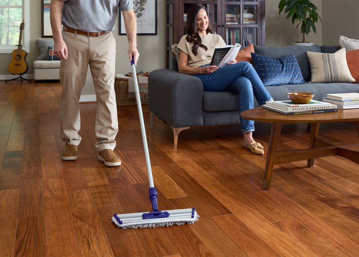 Man cleaning hardwood flooring with a floor cleaning pad while woman sits on sofa